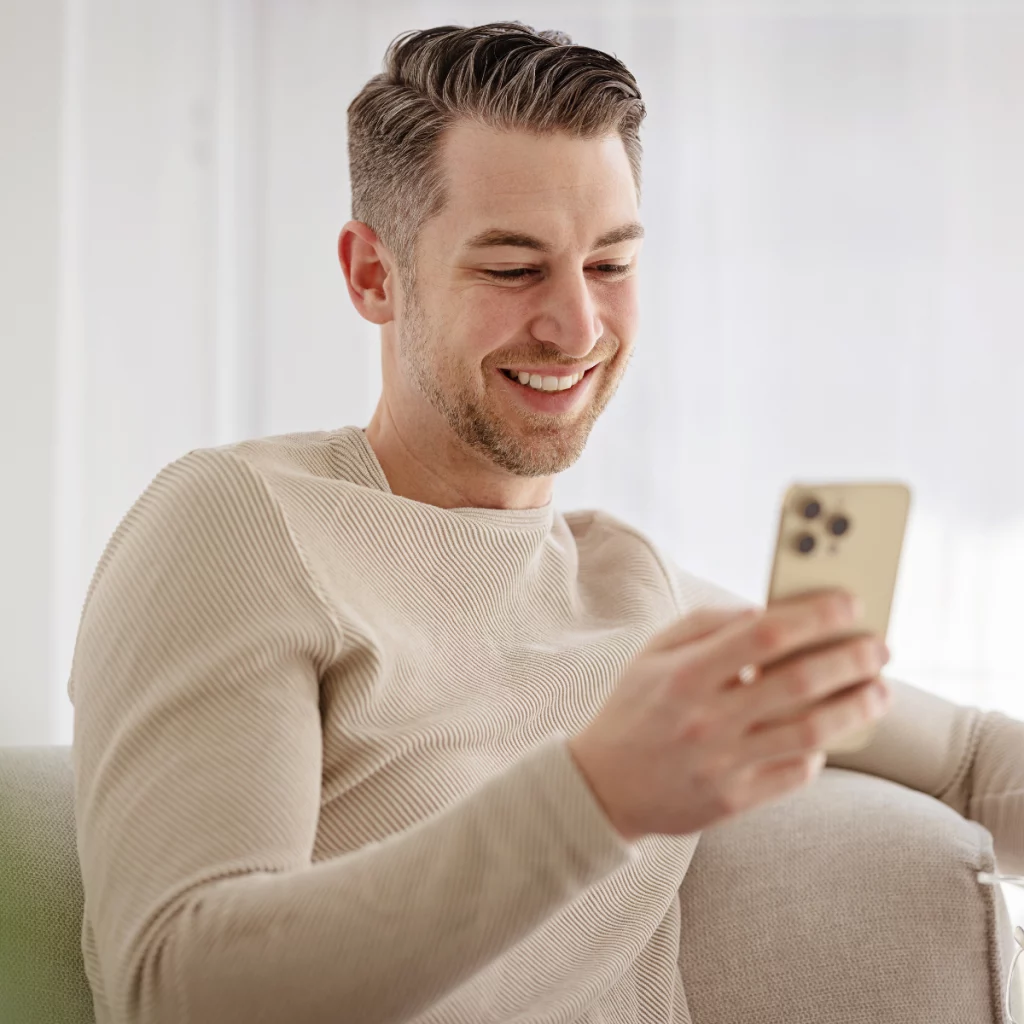Un homme détendu, assis sur un canapé, sourit en consultant son smartphone dans un intérieur lumineux.
